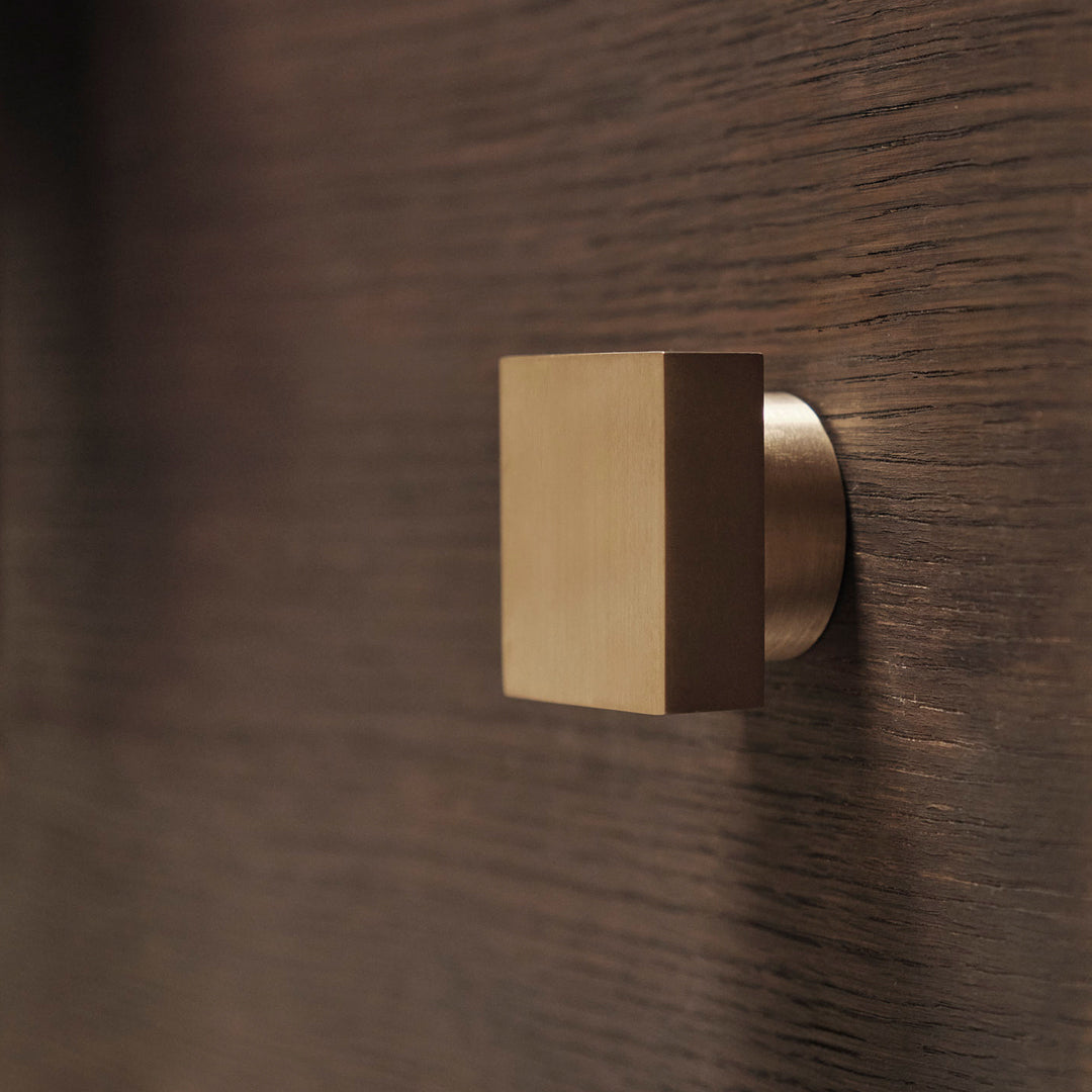 Square brass knob against a dark wood background, showcasing modern minimalist design and textured patterns.