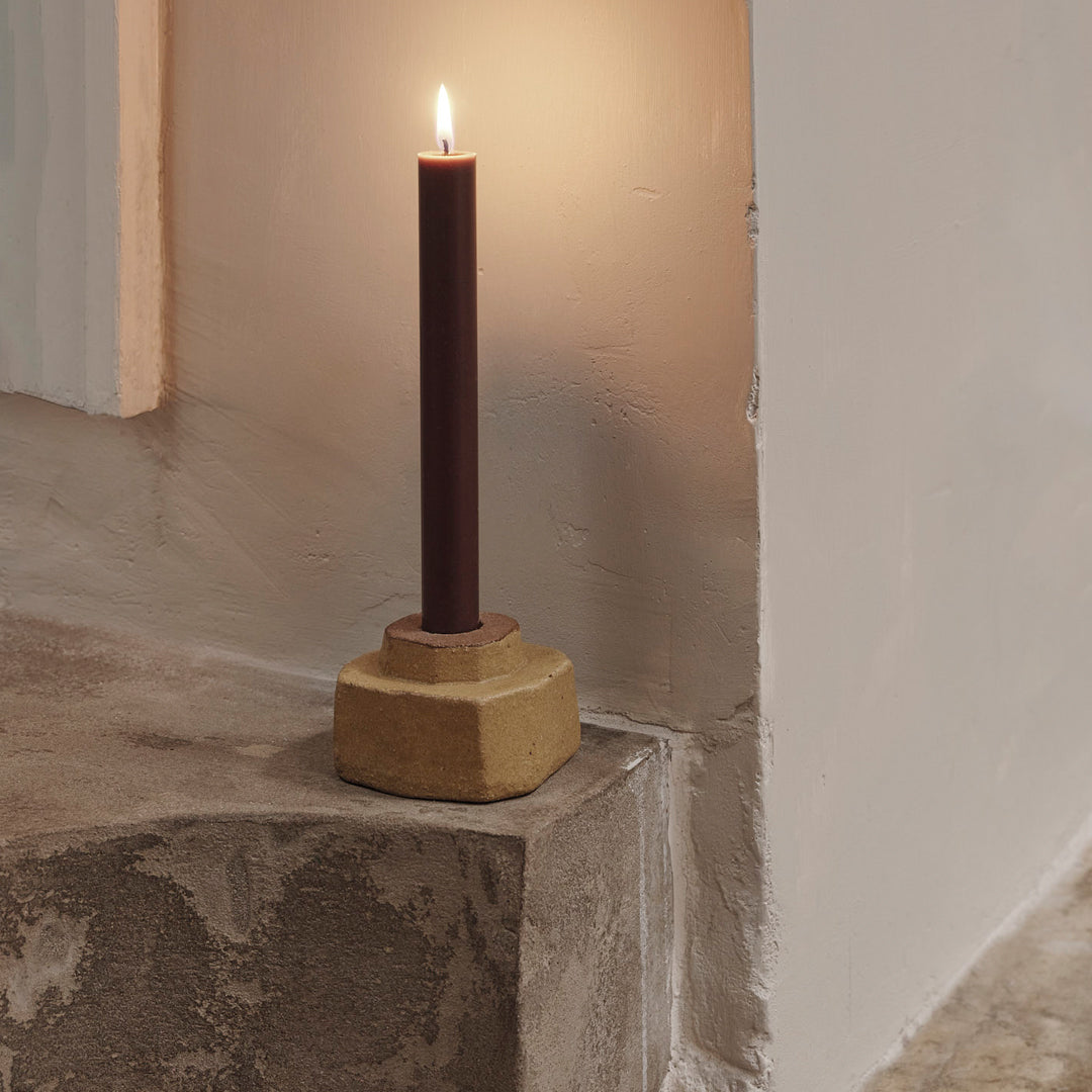 Dark burgundy candle with a textured, neutral-toned holder, set against a soft, light background.