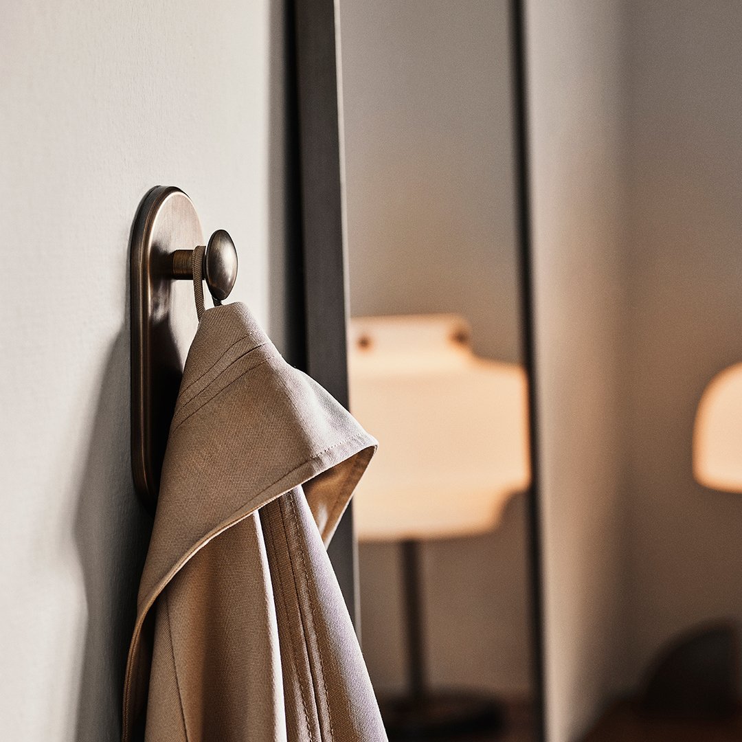 Beige coat draped on a round metal hook against a neutral wall, with soft lighting in the background.