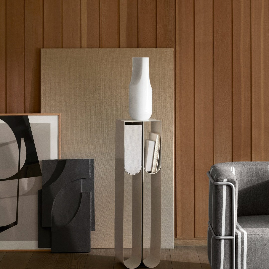 White vase on sleek silver stand, surrounded by black and white abstract art and warm wooden panels. Modern, minimalistic design.