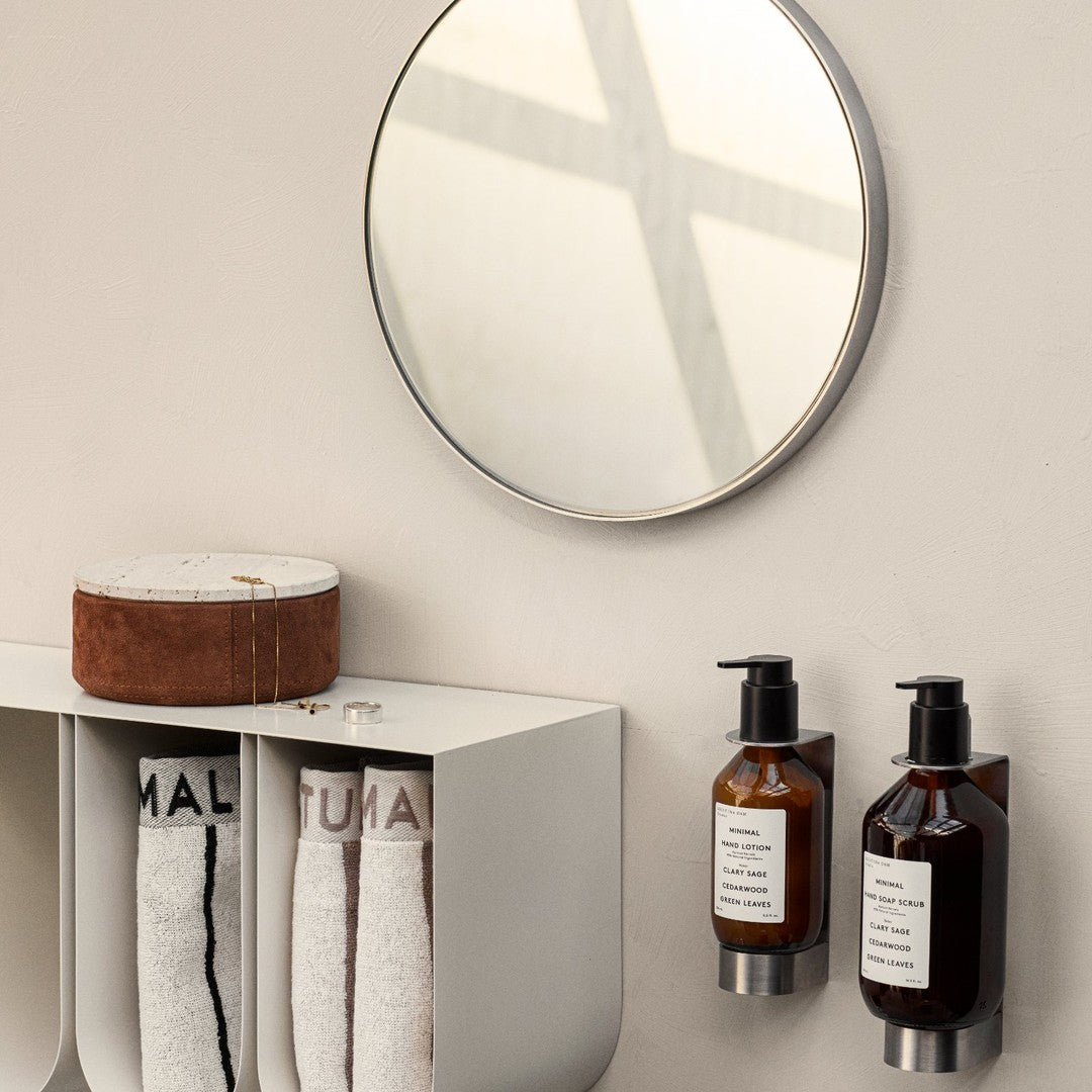 Round mirror reflecting light, black and amber bottles on a white shelf, and white towels with black lettering.