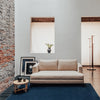 Beige sofa on a dark blue rug against a textured gray and brick wall, complemented by light-toned windows.