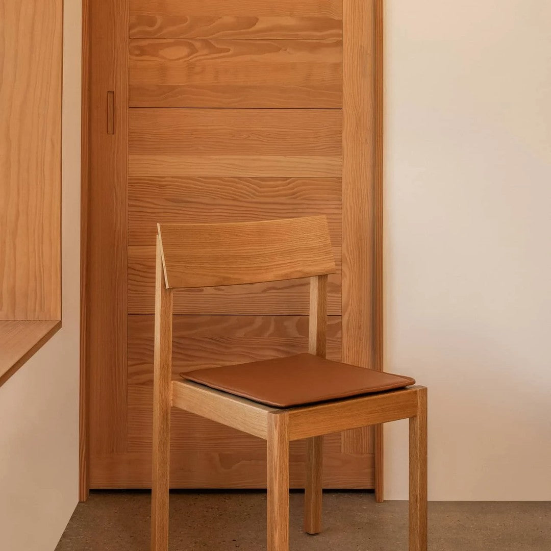 Wooden chair with a natural finish and a warm brown leather seat, set against a neutral beige background.