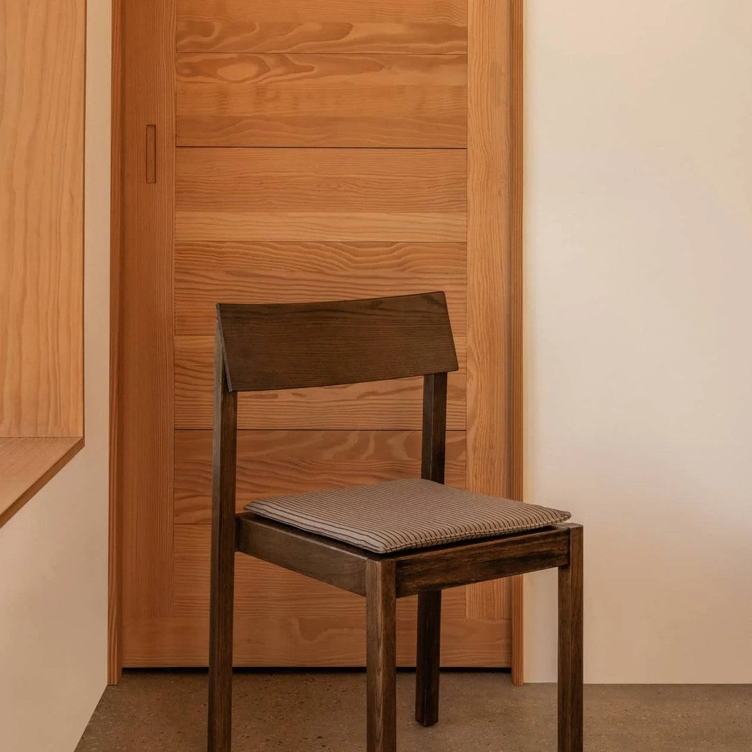Dark wooden chair with a minimalist design and a light gray fabric cushion against a warm wood backdrop.