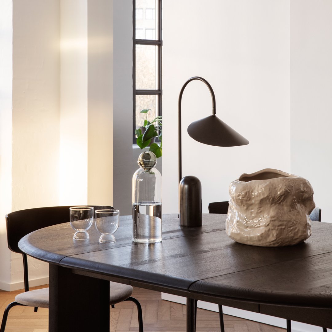 Black round table with a textured white vase, clear glass accents, and modern black lamp, featuring neutral wall tones.