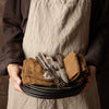 "Black plates stacked with earthy brown cloth napkins and shiny silver utensils, displayed against a warm backdrop."