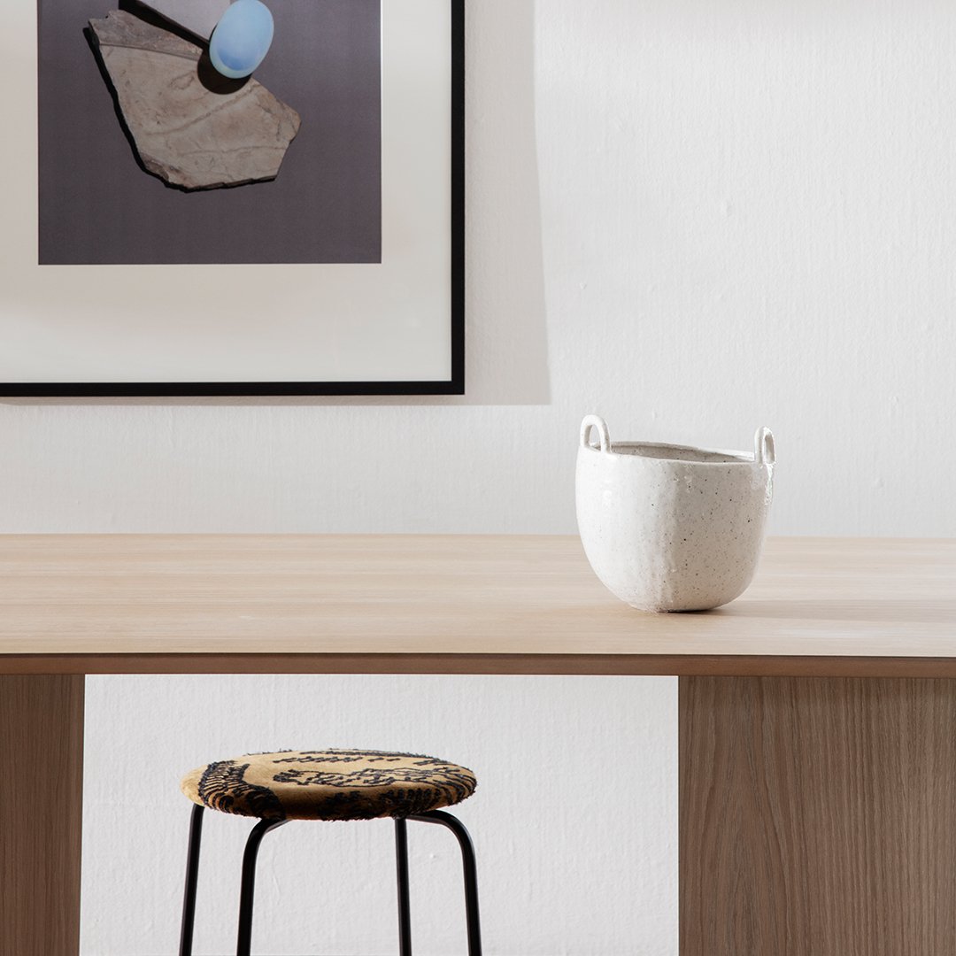 White textured basket on a wooden table, featuring a patterned stool with earthy tones and black legs. Minimalist decor.