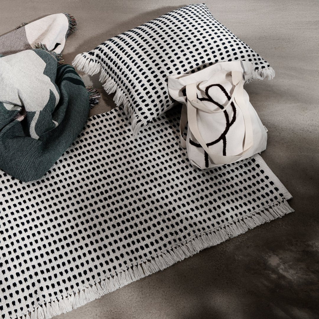 Black and white patterned rug with fringes, paired with patterned pillows and a dark green blanket on a gray surface.