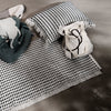 Black and white patterned rug with fringes, paired with patterned pillows and a dark green blanket on a gray surface.