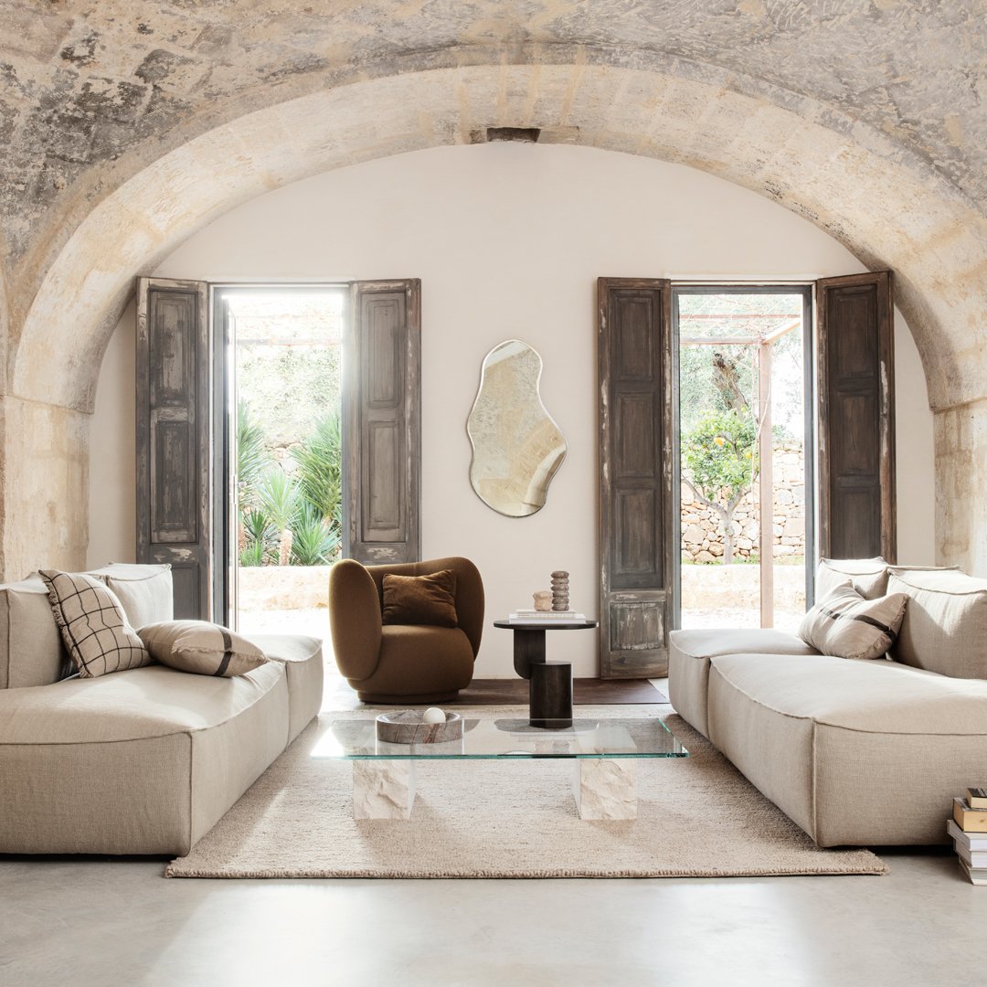 Modern living room featuring neutral beige sofas, a brown accent chair, and natural light from open wooden windows.