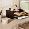 Black wooden organizer with layered shelves, set against a textured beige wall, featuring earthy brown and green accessories.