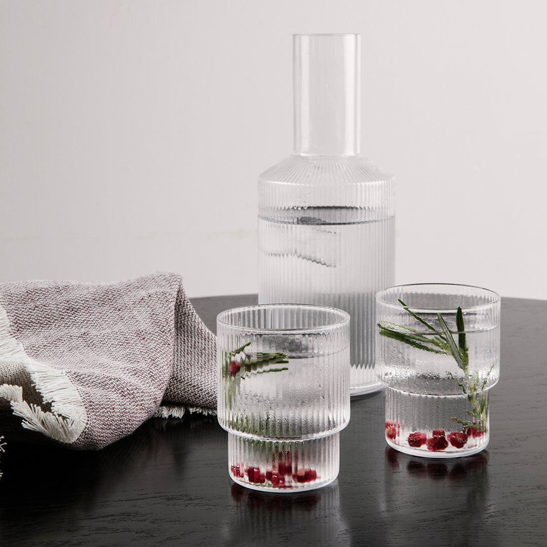 Clear ribbed glass water carafe and tumblers with fresh herbs and berries, set on a dark wooden table with a textured towel.