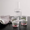 "Clear ribbed glass pitcher and glasses with green herbs and red accents on a wooden table, complemented by a textured cloth."