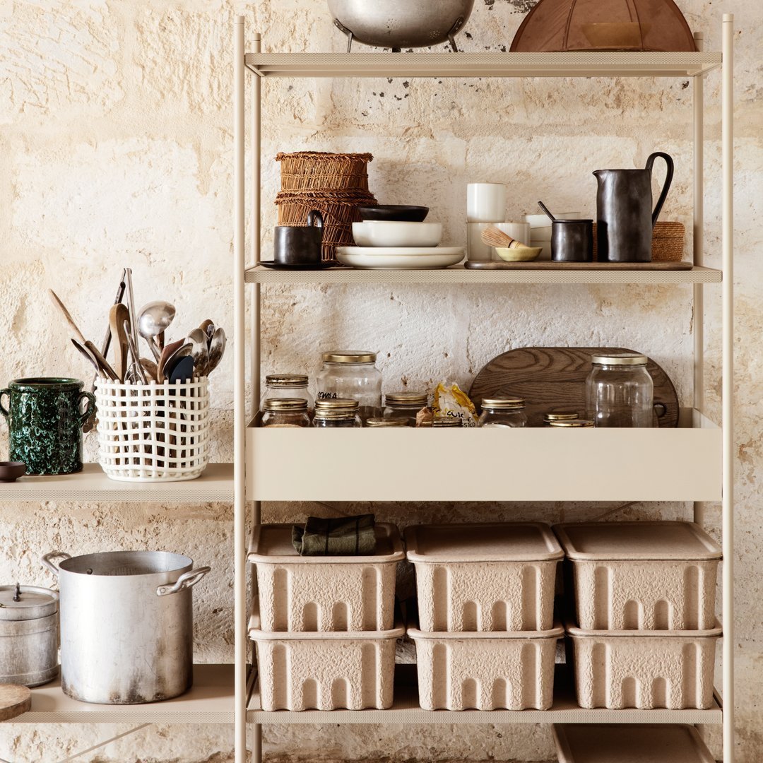 Cream and beige shelving unit with woven baskets, glass jars, and metallic accents against a textured stone wall.