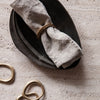 "Neutral gray linen napkin wrapped with a gold ring, set on a matte black ceramic plate, featuring organic shapes."