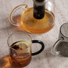 "Clear glass teapot with amber tea and two unique mugs featuring black and gray colors."