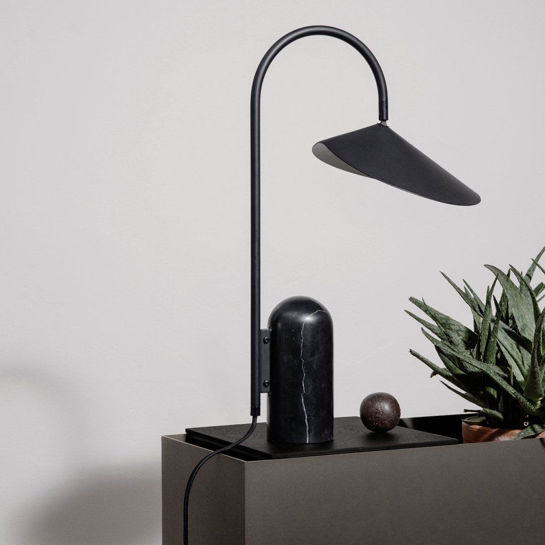 Sleek black lamp with a curved arm and geometric shade, set against a neutral background with green plant accents.