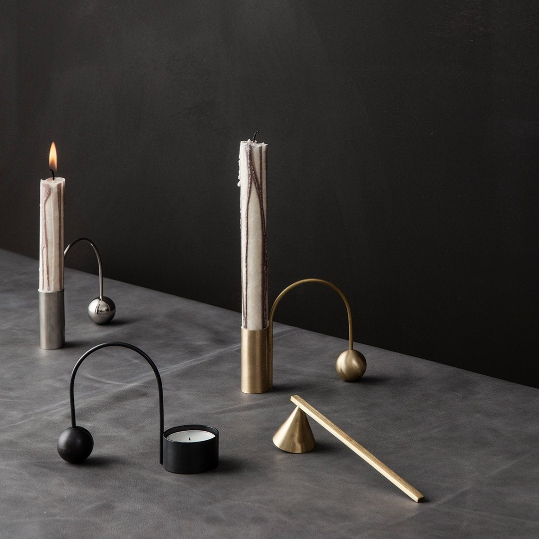 Minimalist candle holders in black, brass, and silver against a dark gray surface, featuring sleek, modern designs.