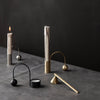 Minimalist candle holders in black, brass, and silver against a dark gray surface, featuring sleek, modern designs.