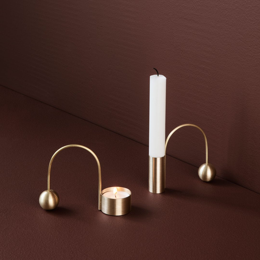 Gold candle holders with curved designs, featuring a white candle and a tea light against a rich brown background.