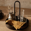 Black circular tray with clear glasses, metal pitcher, silver cutlery, and mustard yellow linen napkin, set on a wooden table.