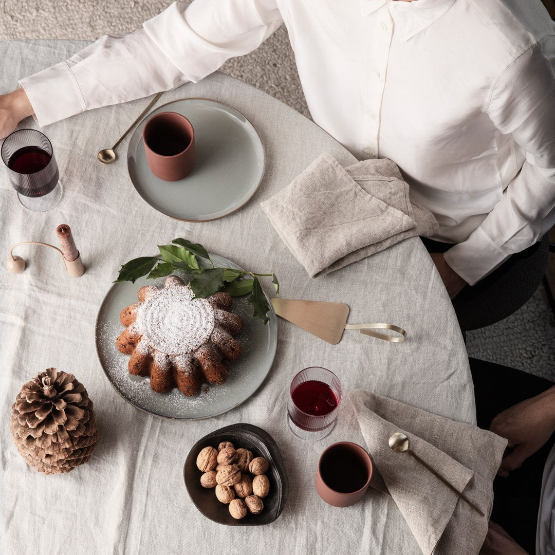 White tablecloth with neutral plates, dark red glasses, and a brown bundt cake surrounded by natural elements.