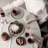 White tablecloth with neutral plates, dark red glasses, and a brown bundt cake surrounded by natural elements.