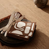 Brown napkins with white abstract patterns on a black plate, featuring silver cutlery, styled on a woven tablecloth.