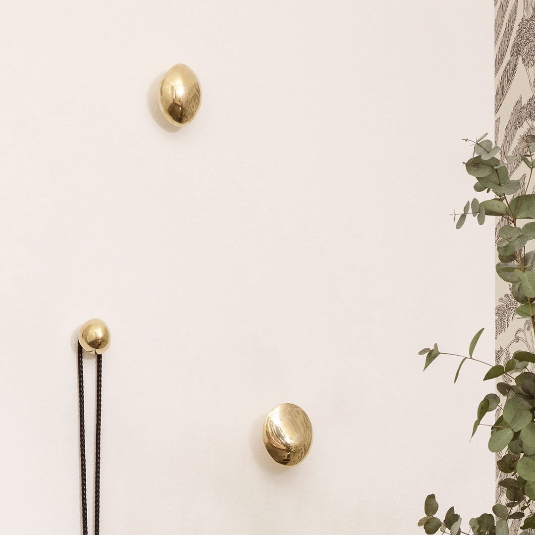 Gold wall hooks with smooth, modern shapes against a neutral wall, complemented by green foliage.