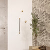 Brass wall hooks against a textured floral wallpaper in gray and white, with a green potted plant.