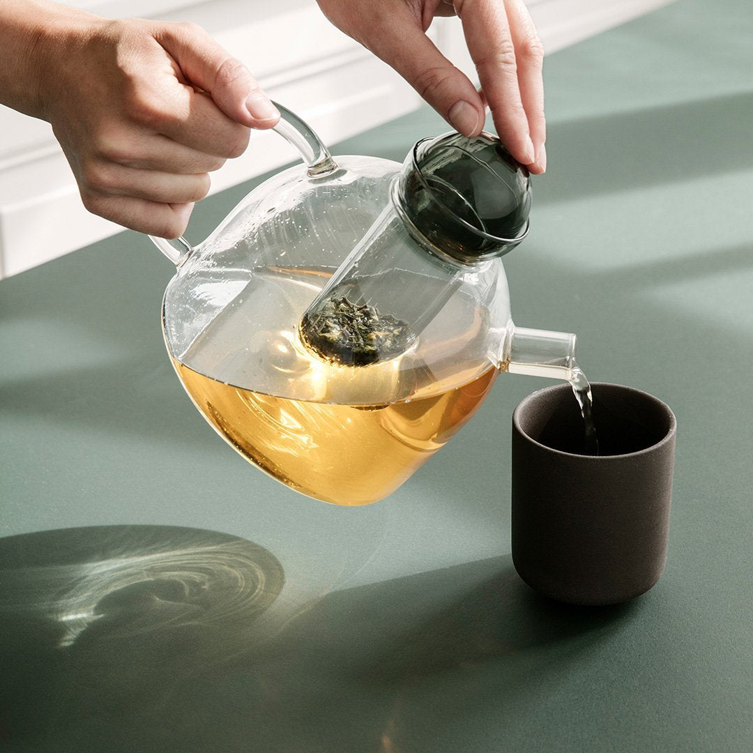 Clear glass teapot pouring golden tea into a dark brown cup, set against a soft green background.