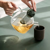 Clear glass teapot pouring golden tea into a dark brown cup, set against a soft green background.