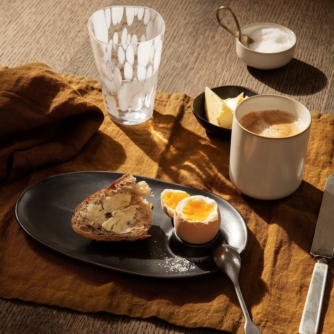 White patterned glass, dark oval plate, warm brown cloth, neutral ceramic cup, and creamy soft-boiled egg on a textured surface.