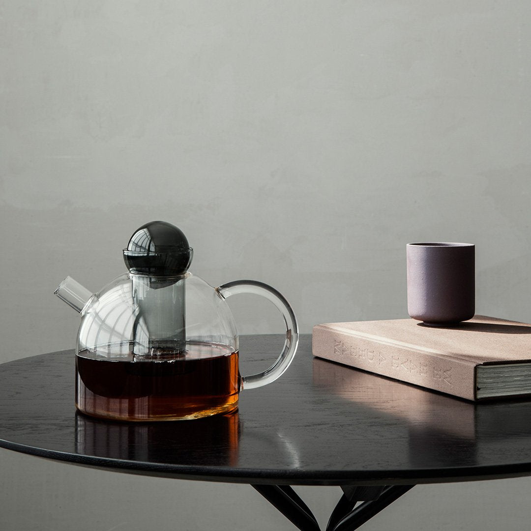 Clear glass teapot with black lid, amber tea, and a minimalist purple cup on a sleek black table.