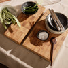Wooden cutting boards with a warm checkerboard pattern, black ceramic bowls, and natural green vegetables on a light tablecloth.