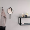 Round mirror with a sleek black frame against a muted gray wall; stylish striped towel in soft pink and black. 