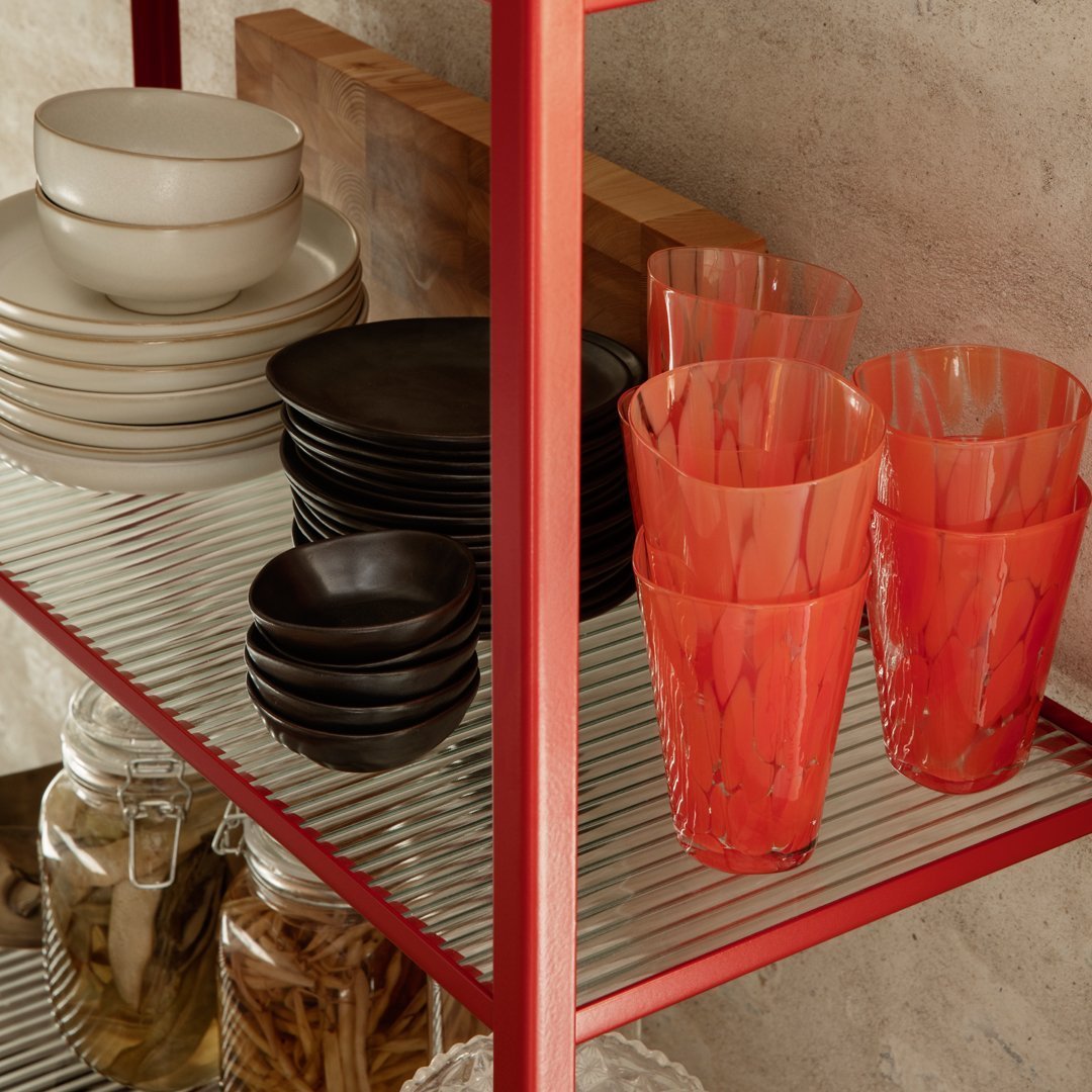 "Colorful glassware in vibrant orange and black, arranged on a red metal shelf with neutral dishware and jars."
