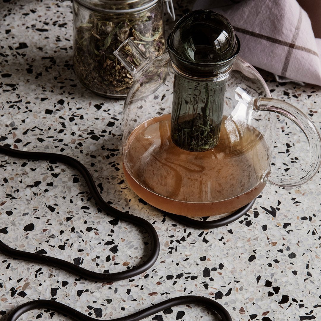 Glass teapot on speckled terrazzo surface with earthy tones and a curved black tea infuser.