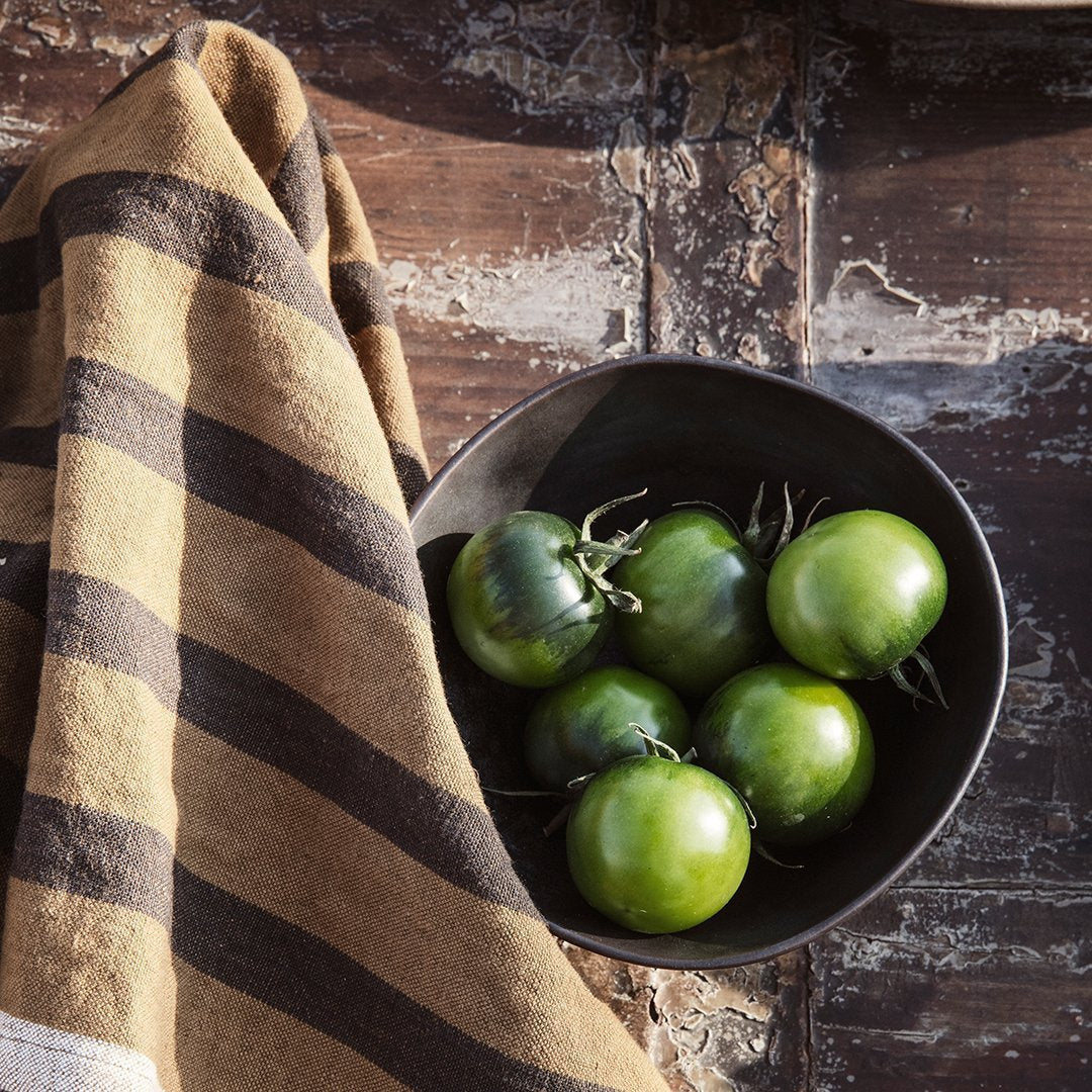 Green tomatoes in a black bowl, contrasted with a striped brown and cream towel on a rustic wooden surface.