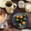 "Round black plate with green and orange tomatoes, rustic ceramic pitcher, and assorted dishware on a wooden table."