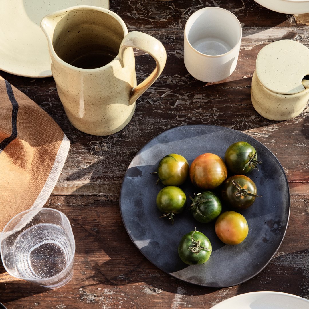 Rustic table setting with earthy tones, green and yellow tomatoes on a dark plate, surrounded by minimalist ceramics.