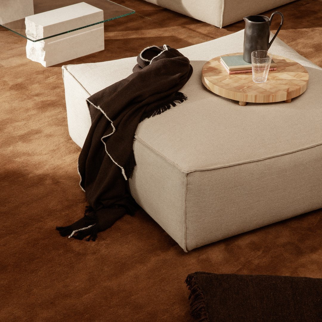 Neutral beige pouf with dark brown throw, set on a rich brown carpet, complemented by a wooden tray and glass.