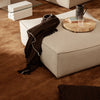 Neutral beige pouf with dark brown throw, set on a rich brown carpet, complemented by a wooden tray and glass.