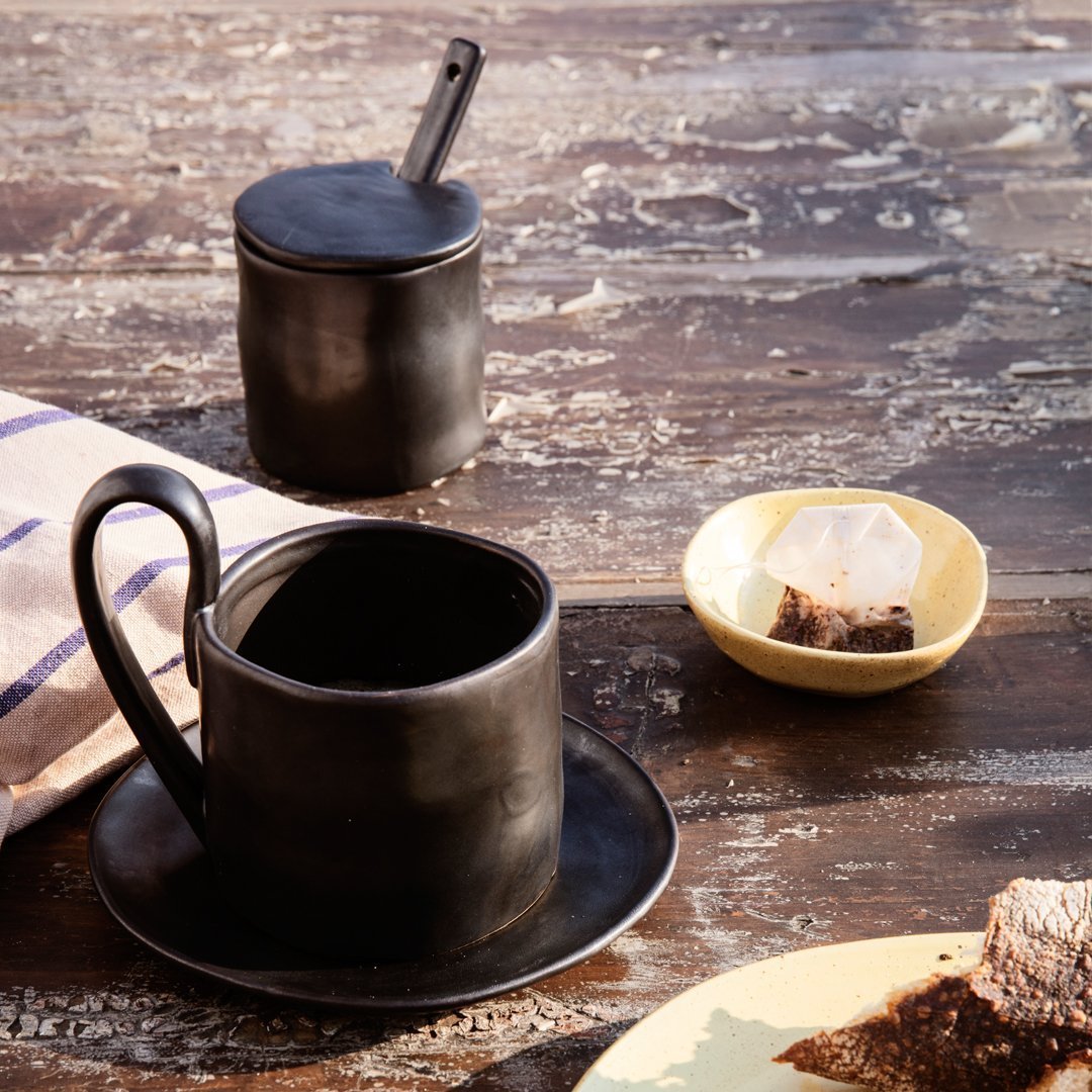 Black ceramic cup and saucer set with a matte finish, accompanied by a sleek black sugar container and a yellow dish.