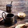 Black ceramic cup and saucer set with a matte finish, accompanied by a sleek black sugar container and a yellow dish.