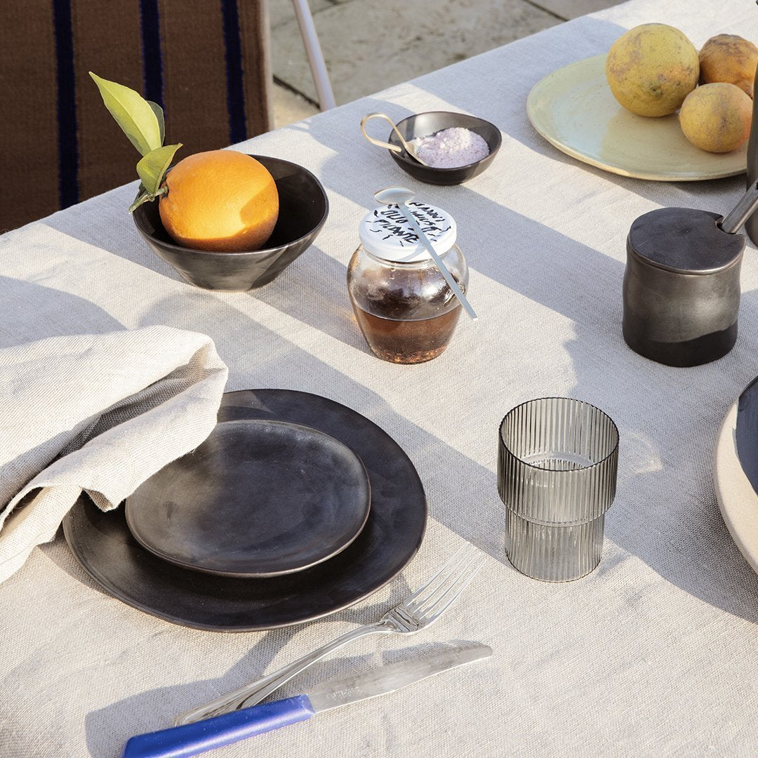 Black ceramic plates, textured glass cups, and neutral linen tablecloth create a modern, stylish dining setting.