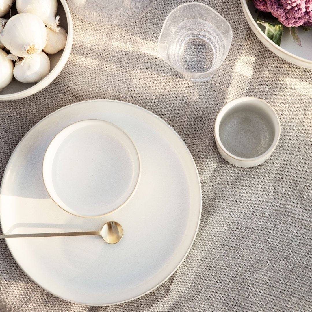 Neutral-toned dining set featuring smooth ceramic plates, a glass, and minimalist designs on a textured linen tablecloth.