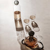 "Clear glass water carafe and cups with amber liquid, casting soft shadows on a neutral surface."