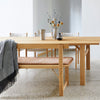 Wooden dining table with a natural finish, beige bench cushion, and grey throw, complemented by soft textured patterns.
