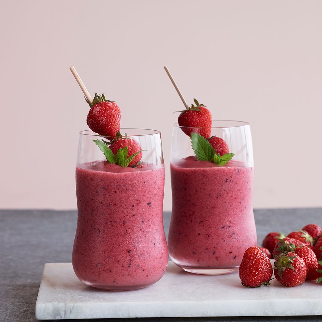 Pink strawberry smoothies in clear glasses, garnished with fresh strawberries and mint leaves, on a marble surface.